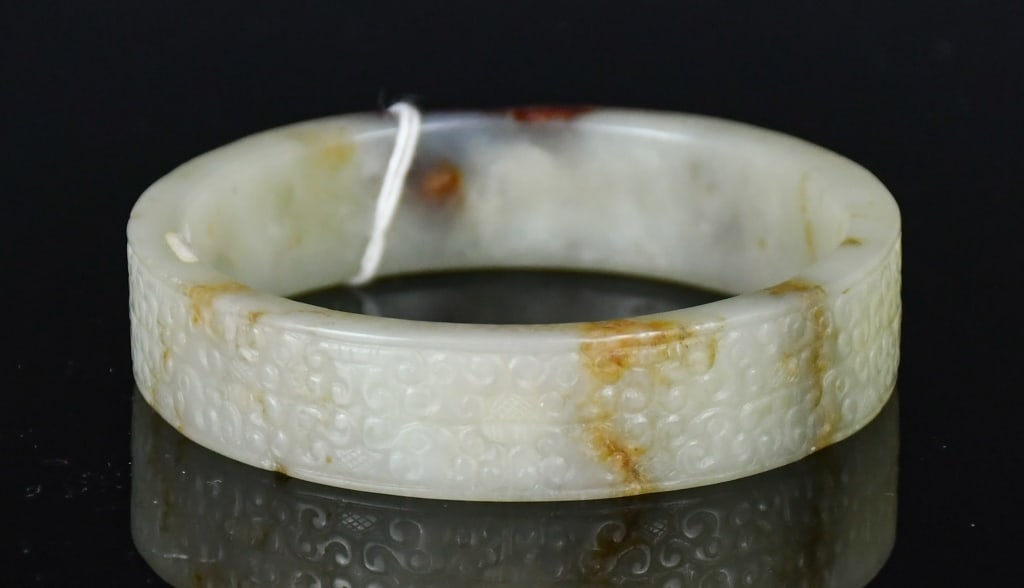 A Jade Carved Bangle w/Box: A jade bangle carved in relief with scrolling motifs around the exterior, the stone of white-celadon tone with russet inclusions. D: 5.6cm 青白玉雕雲紋手Ż