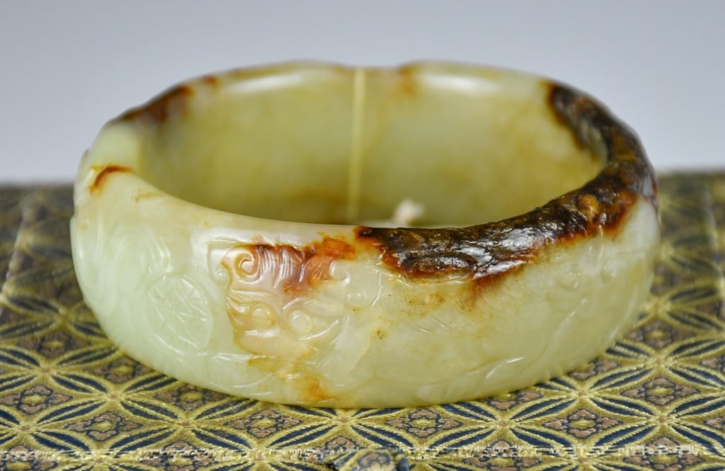 A Jade Carved Bangle w/Box: A jade bangle carved in relief with beast motif patterns around the exterior, the stone of yellowish celadon tone with dark brown and russet inclusions. D: 6.5cm 玉雕獸紋৔