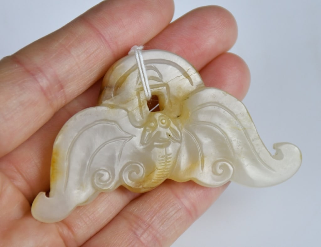 A Jade Carved Pendant: Modelled as a bat grasping a coin, the stone of creamy-white tone with minor russet inclusions. 6.5x3.2cm 玉雕福在眼前珮