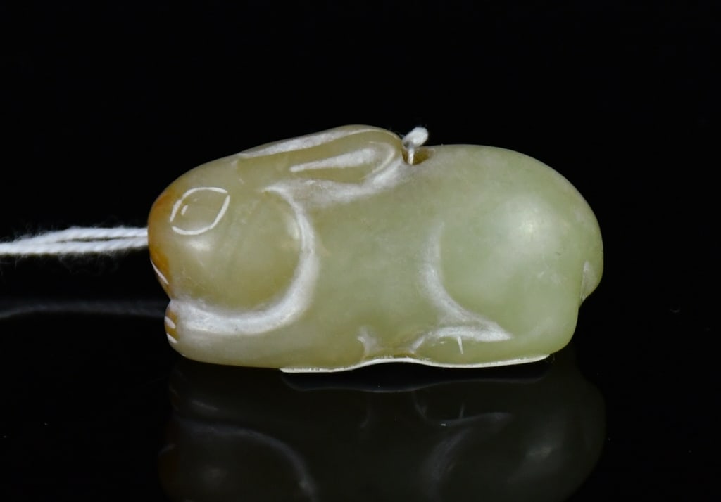 A Celadon Jade Carved Rabbit: The celadon jade carving modelled as a recumbent rabbit with ears laid back along the body, the stone with russet inclusions. 3.3x1.2cm 青玉雕臥兔擺件