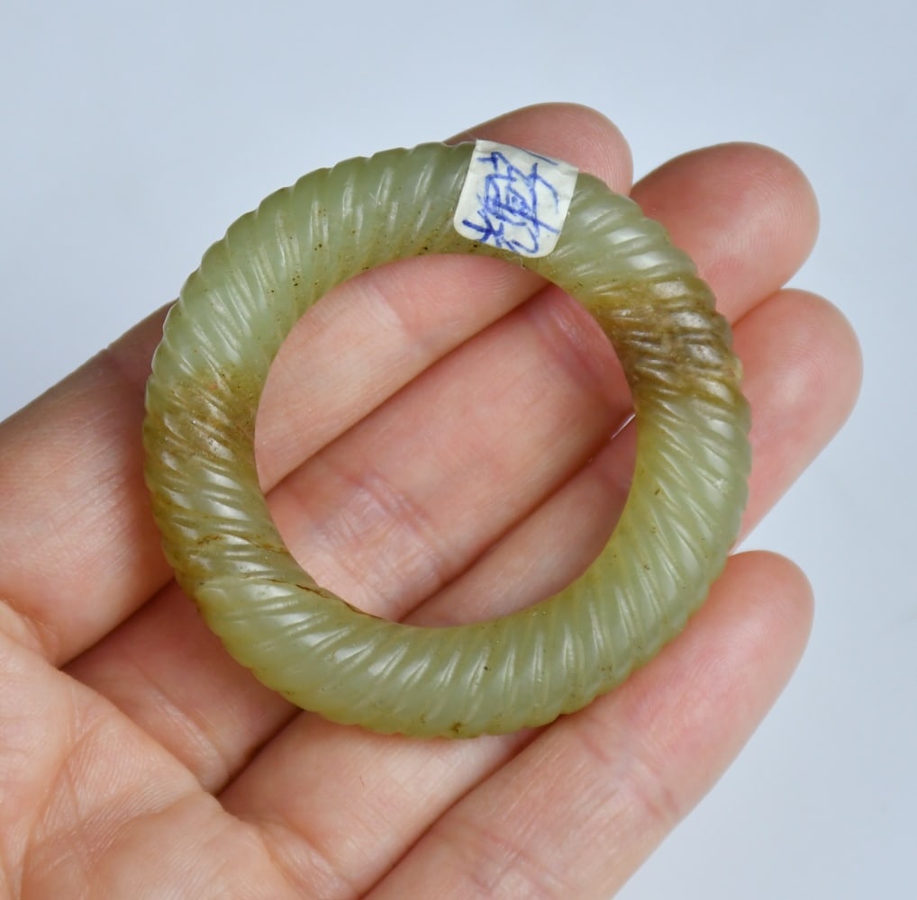 Archaistic Style A Celadon Jade Carved Huan Ring: A small celadon jade ring of circular form, carved around the exterior with twisted rope patterns, the stone with russet inclusions. Inner diameter: 2.9cm 高古風格青ħ