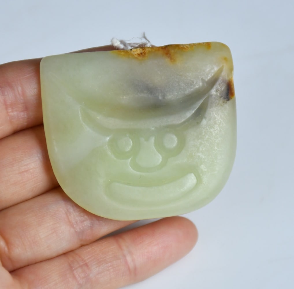 Archstic style A Celadon Jade Carved Mask Plaque: A celadon jade plaque of rounded rectangular form, carved in low relief with mask motif, with russet inclusions. 4.6x4cm 高古風格青玉雕獸面牌