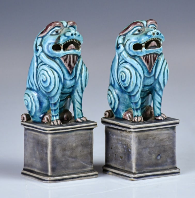 A Pair of Glazed Fo Lions 19thC: Each mythical beast is modeled seated upright atop a tall rectangular plinth base. The creatures are crisply sculpted with bulging eyes, stylized spiraled fur motifs across the body, and an open, holl