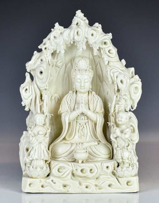 A Blanc-de-Chine Figure of Guanyin & Acolytes Qing: The figure is finely modeled depicting Guanyin, set within a rocky grotto or mountain throne with stylized bamboo and swirling cloud-like formations. Flanking the central figure are two standing