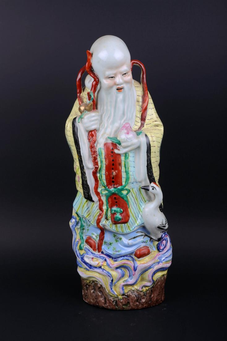 A Famille Rose Shou Lao Figure: A famille rose figure, depicting Shou Lao dressed in a flowing robe, holding his staff hung with a gourd in his right hand and a peach in his left hand, standing on swirling clouds with a crane by his