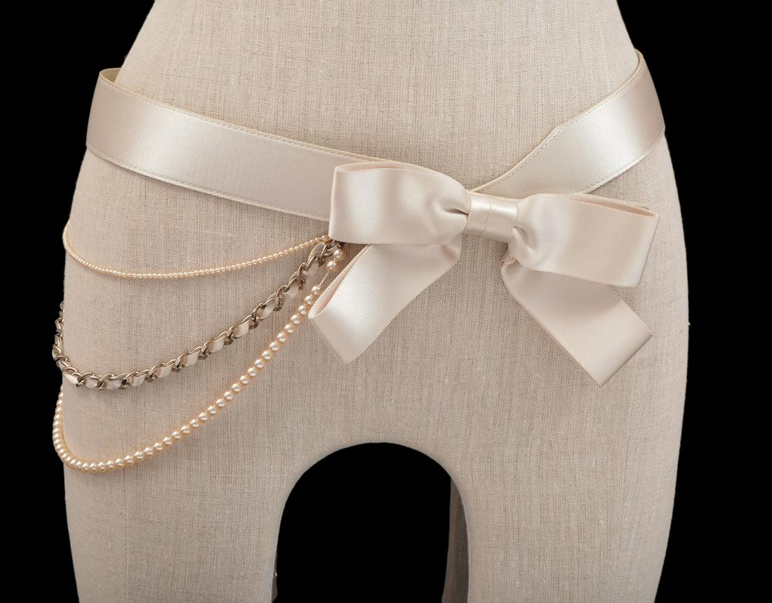 Chanel Beige Satin Bow & Pearl Chain Belt 2002: This beige satin belt has a double strand of faux pearls and chain link interspersed with satin ribbon. It is part of the 2002 Cruise Collection. It features a beige bow covering a 3 snap closure. The