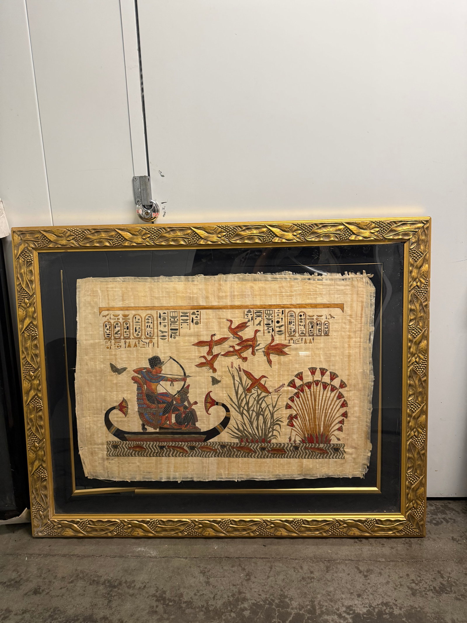 Framed Egyptian Papyrus Art 50”x40”: Hand-painted Egyptian papyrus art is set within a decorative gold-toned frame. This piece has a black mat and features traditional Egyptian motifs including a pharaoh on a boat, birds, and hieroglyphi