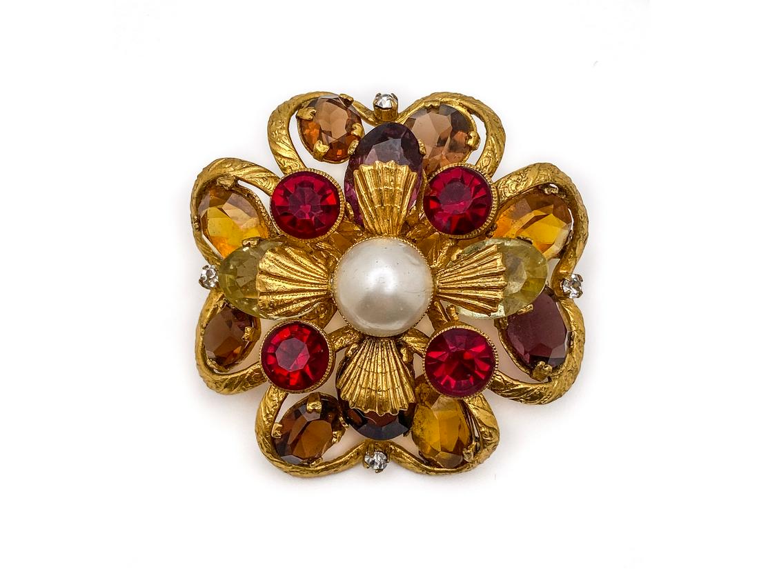 Chanel Pearl and Rhinestones Brooch.: Designed as flower with a central cross motif. Openwork in gilt metal, central large faux pearl with four ruby rhinestones, topaz, and smoky rhinestones, completes the petals design. Oval tag on the b
