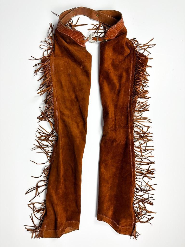 Amber Suede Chaps with Sterling Hardware Branson Colorado Leather: Sterling buckle stamped K / J. (2) silver 2 in. dia. conchos. Measures 12 in. at top of leg when laying flat. 48 in. from top of waist to bottom hem. Weight 3 lbs. Provenance: From the Estate of Nick