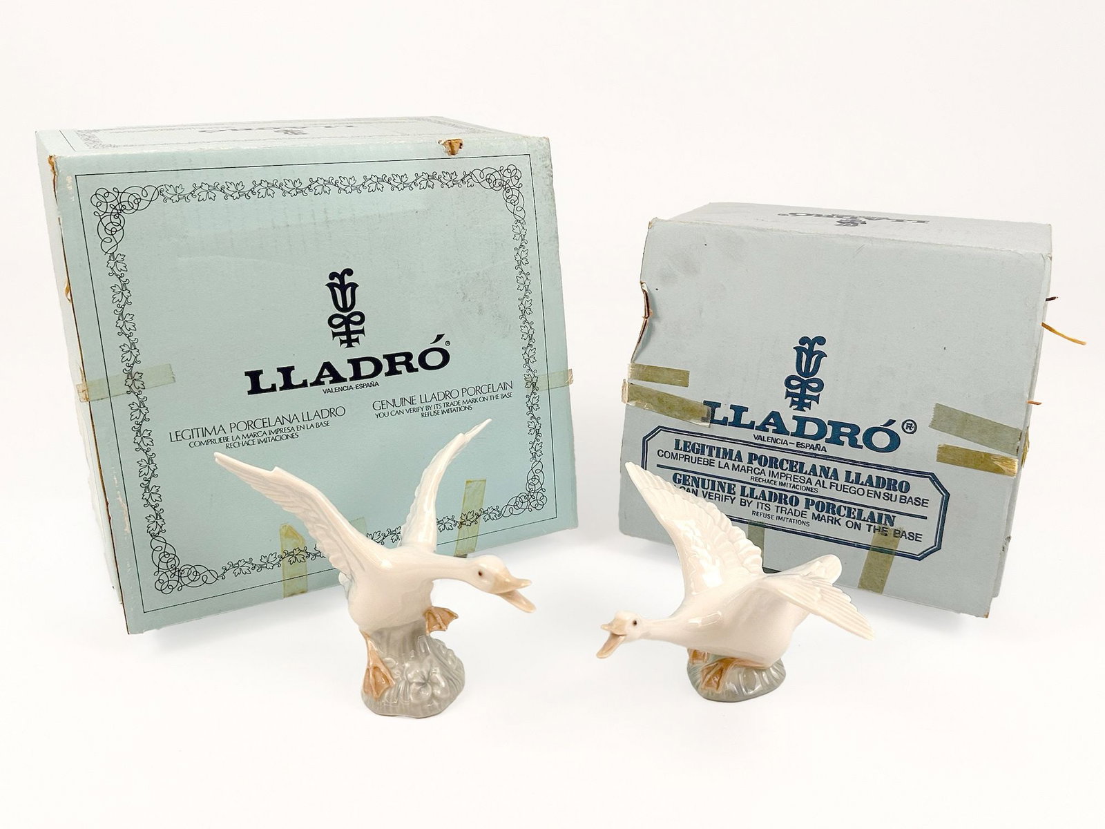Lot of 2 of Lladro Porcelain Goose Figurines with Original Boxes: Pair of porcelain goose figurines, each depicted with wings spread and open beaks. The figurines are mounted on small, stylized bases. Each comes with original box. Each sculpture is approximately 4.5
