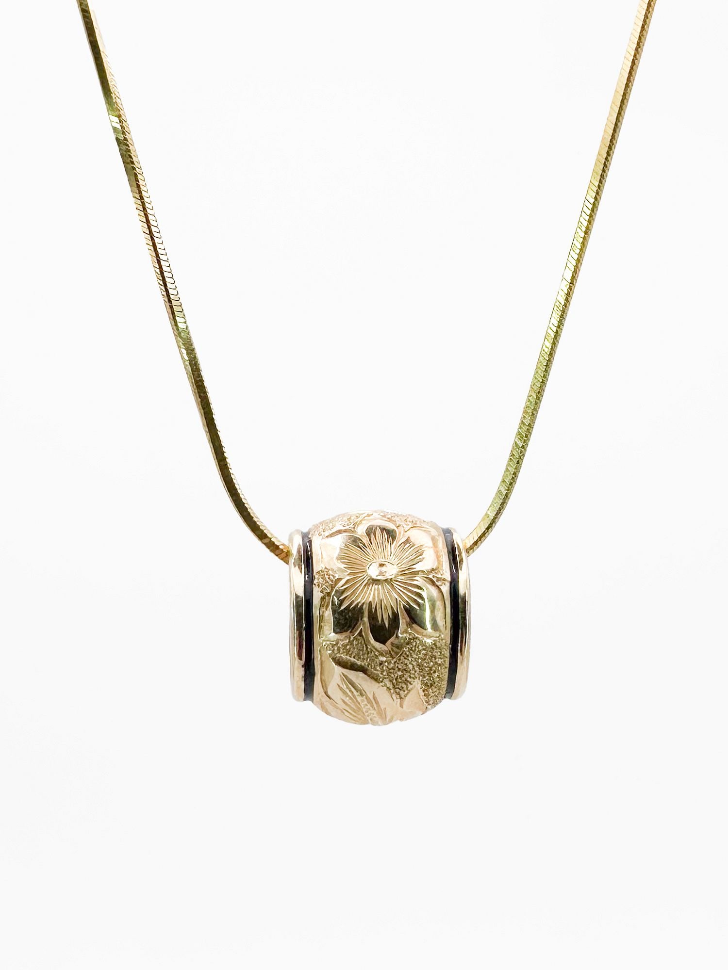 14K Gold Plumeria Scroll Puka Pendant Necklace: 14K yellow gold puka pendant with black enamel borders and a plumeria scroll design. The pendant measures approximately 3/8 inch in width. Includes a 16-inch 14K snake chain. Total weight: 9.6 grams.