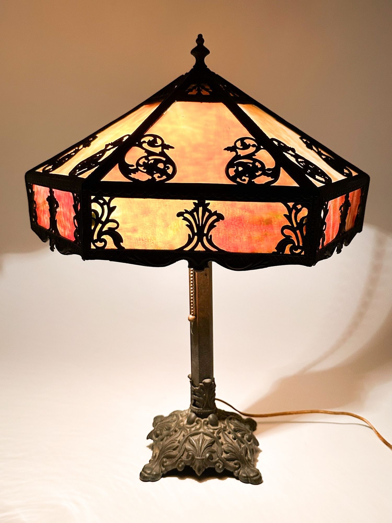 Antique Slag Glass Table Lamp with Ornate Metal Base: Lamp is constructed of metal and stained glass. Ornate cast metal features traditional fleur and tassel embellishments. Stained glass is in shades of green, red, and peach. No markings. Approximately 