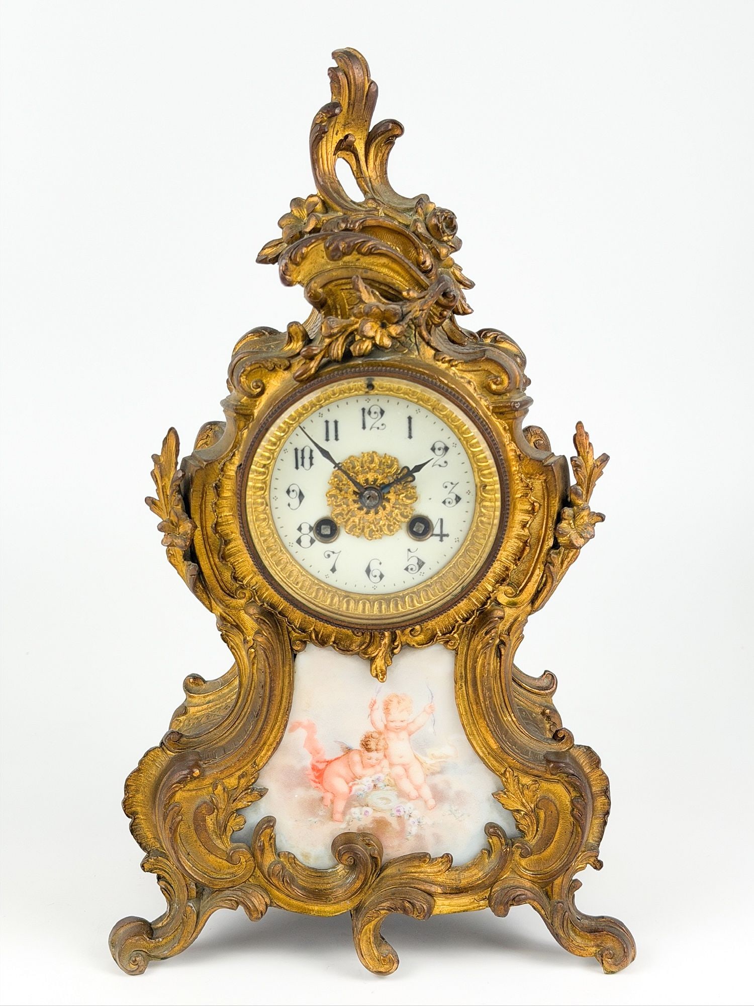 Antique Gilt-Bronze Mantel Clock with Ceramic Cherub Motif: French Rococo-style mantel clock in cast gilt metal, with elaborately scrolled and foliate case, white enamel dial with Arabic numerals and a gilt rosette center. Made of gilt-bronze with ceramic cher