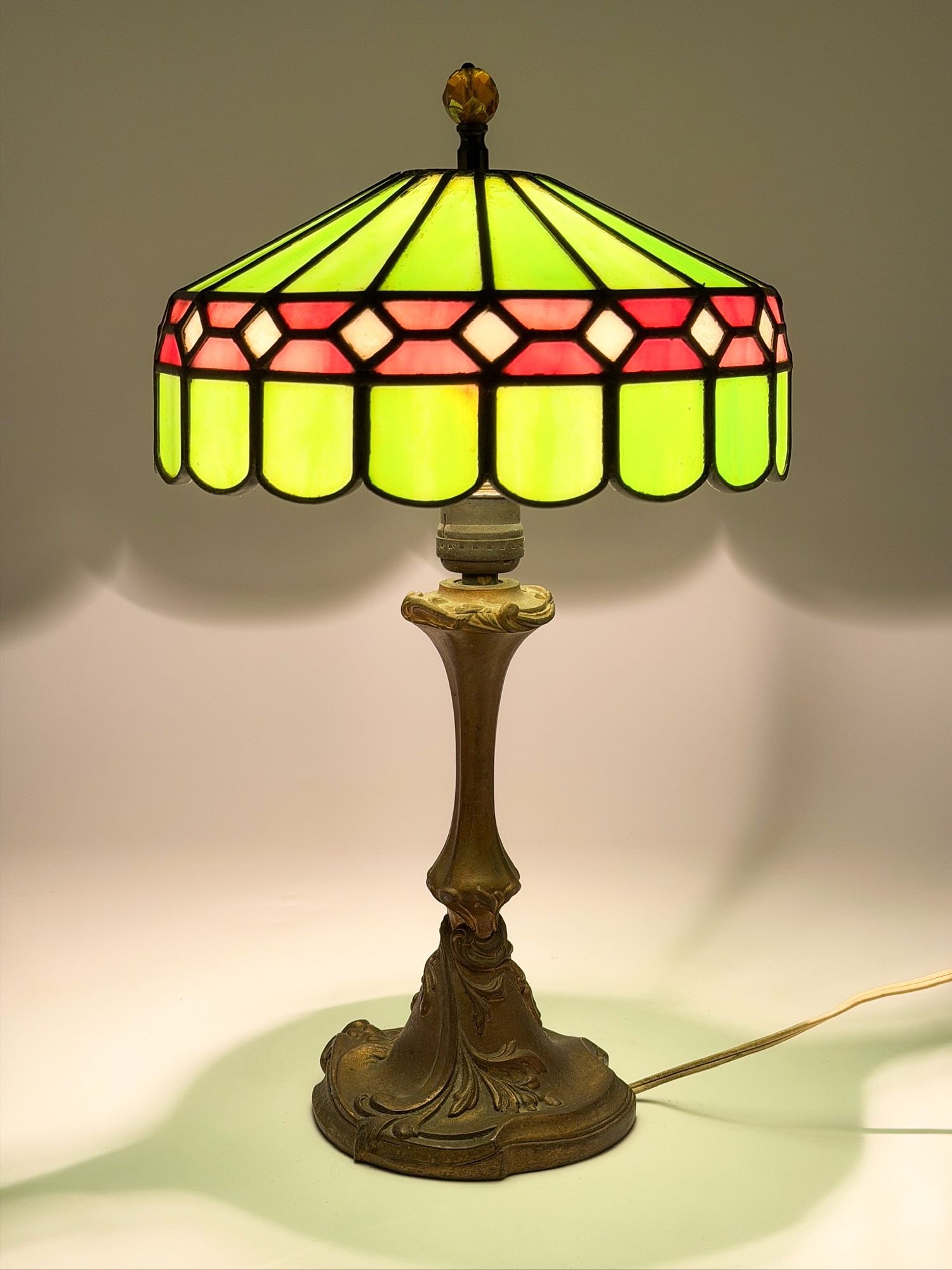 Leaded Stained Glass Table Lamp on Cast Base, Tiffany Style: Lamp features slag glass lampshade and cast metal base. Glass panels come in shades of green, pink, and white. Orange cut glass finial. Approximately 16 in H x 9.5 in W x 9.5 in D. Weighs approximatel