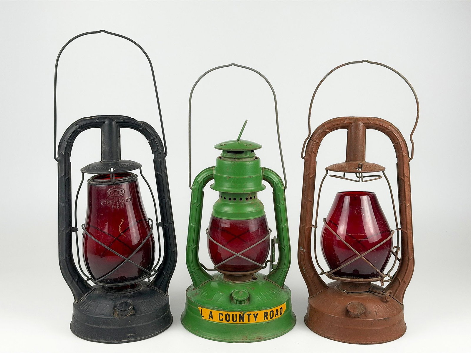 Lot of Three Dietz Res Glass Antique Lanterns: Varying models, two monarch and one No. 100. Constructed of metal and red glass. Sizes range between 13.25 in H x 7.25 in W x 7.25 in D and 13.5 in H x 7.5 in W x 6.25 in D with handles lowered. Total