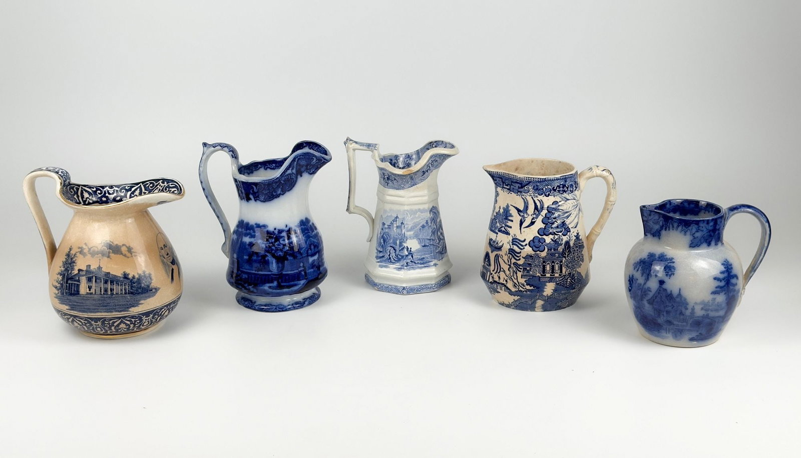 Lot of Five Blue & White Transferware Pitchers: Includes Buffalo Pottery "Mount Vernon"/Washington Jug, Flow Blue, and Willow Pattern. Lot includes pitchers of varying size and shape. Porcelain depicts various scenes of landscapes and portraits. Si