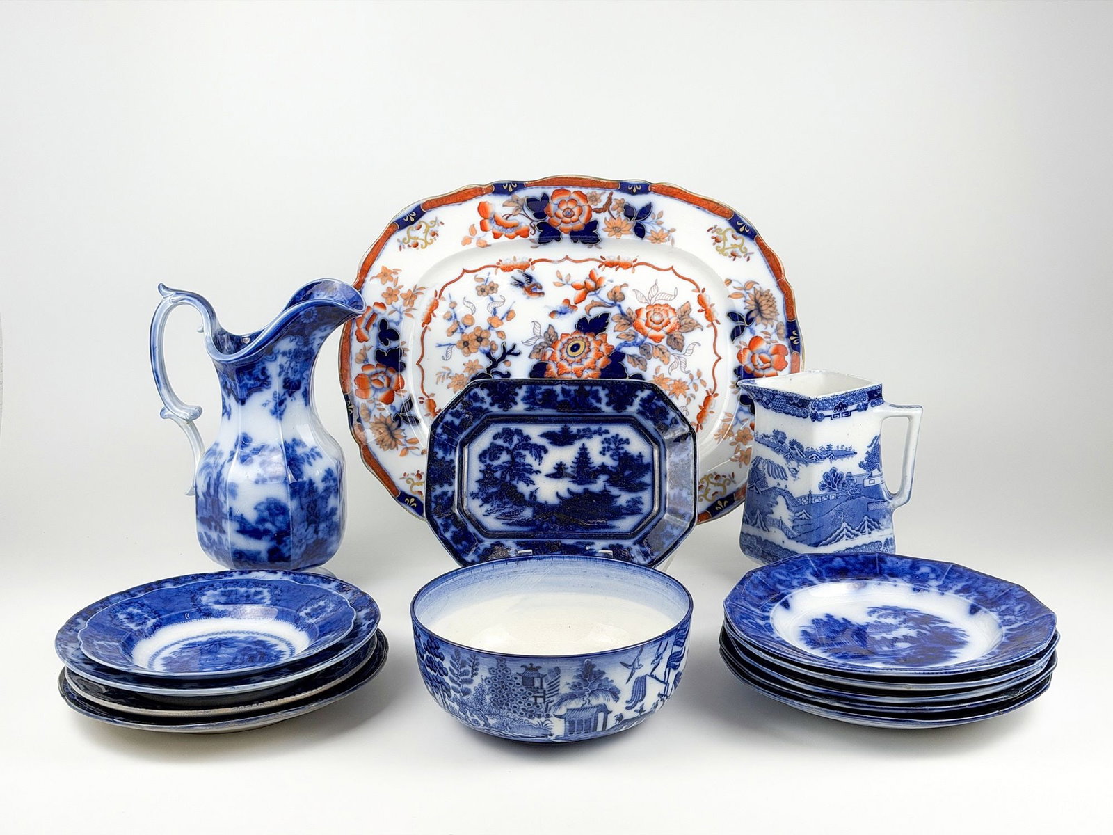 Lot of 14 Victorian Flow Blue & Transferware: Nine Plates Plus Imari Palette Platter, Flow Blue Jugs, Willow Pieces & Ringtons Caddy. Lot includes one serving platter, two pitchers, a collection of 9 plates, one bowl, and one rectangular plat