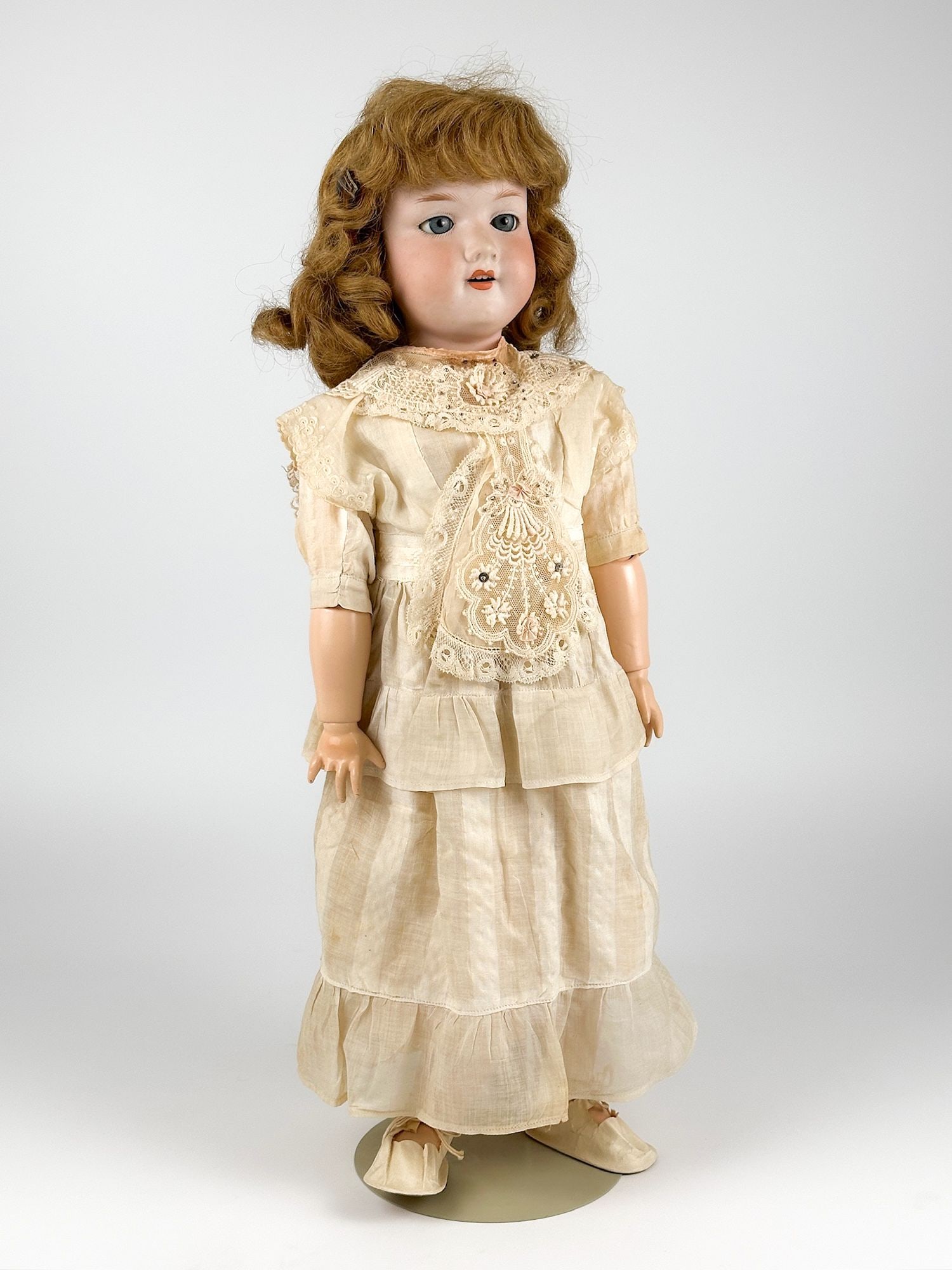 Antique Armand Marseille Porcelain Doll: Bisque porcelain with articulated joints. Dress is linen with lace collar and bib. Made in Germany. Engraved on the back of the neck. Includes stand. Approximately 22 in H x 7.5 in W x 6 in D. Weighs 