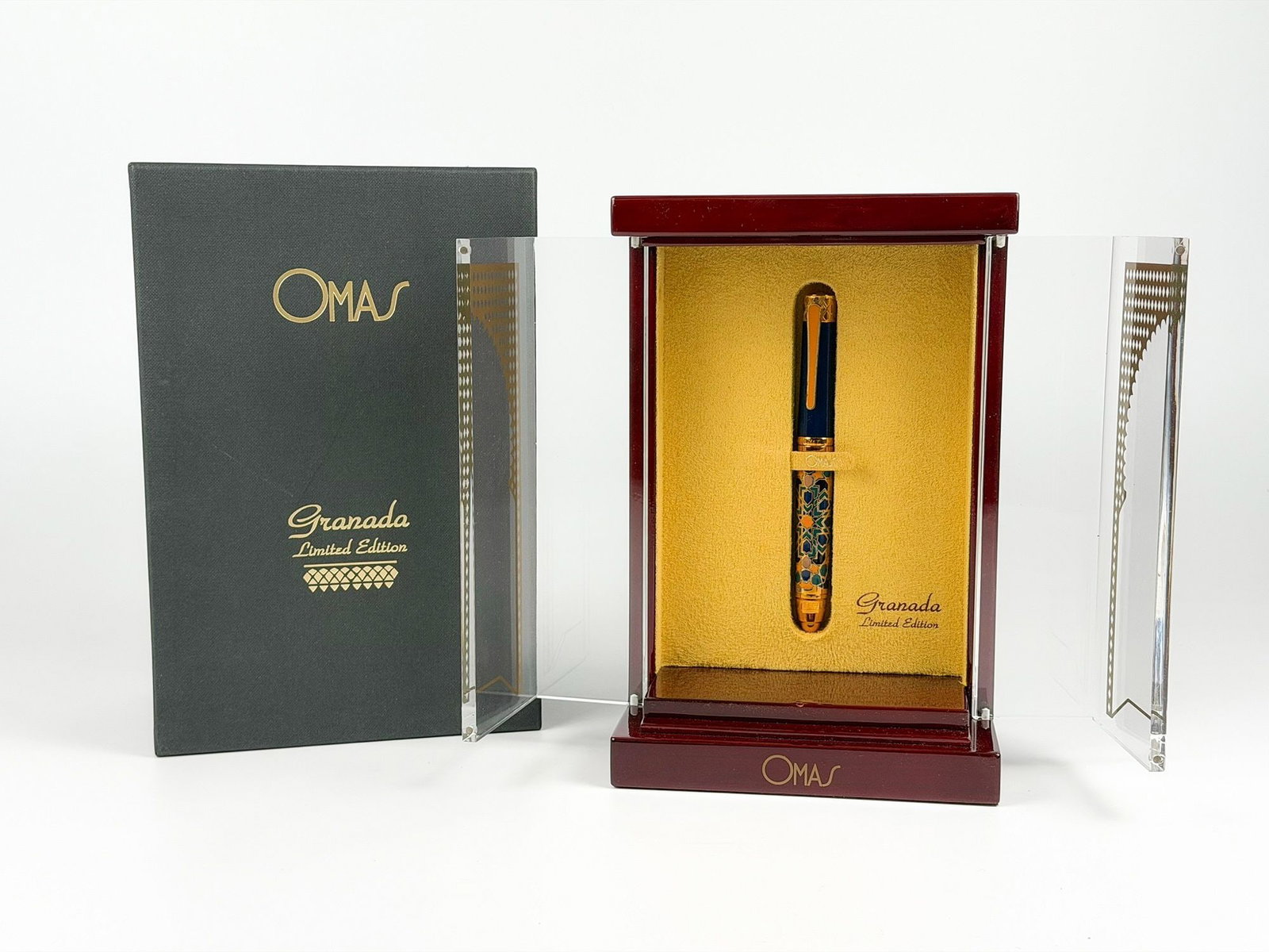 OMAS Granada Limited Edition Fountain Pen in Display Case: A limited edition OMAS fountain pen from the Granada collection. The pen features a dark blue barrel with intricate gold and colorful detailing. It is presented in its original wooden display case wit