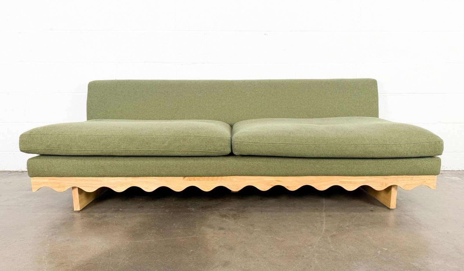 Scalloped Wood Sage Green Sofa: Sleek and whimsical design with no arm rests and linen textured upholstery in a sage green color. Wavy bottom wooden trim wraps around the whole sofa. Dimensions: Approx dimensions: 25 in H x 75 in W 