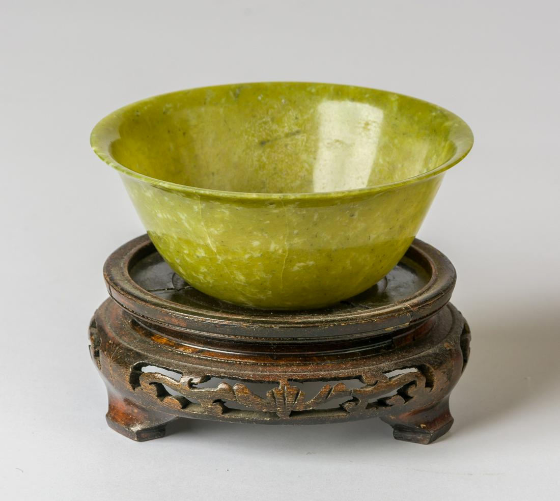 A GRRENISH BOWL: A GRRENISH BOWL China, Jade, probably 19th c. 12.9 x 6 x 12.9 cm, wooden base: 12.9 x 5.3 x 12.9 cm Condition: The bowl has a crack.
