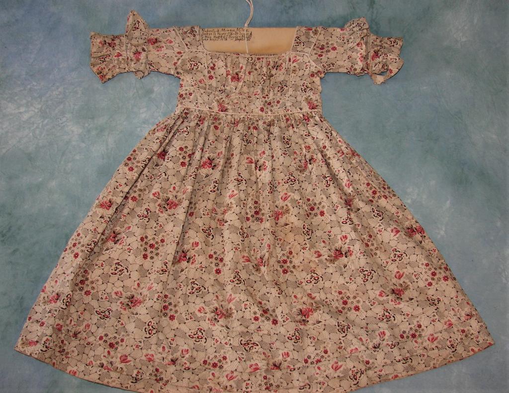 Regency Era Printed Cotton Dress, 1835: Small dress for a child on a cream cotton that is printed with allover with floral and abstract images in reds, pinks, blacks, browns. Ruffled sleeves, pleated bodice and waistline, piping, three flou