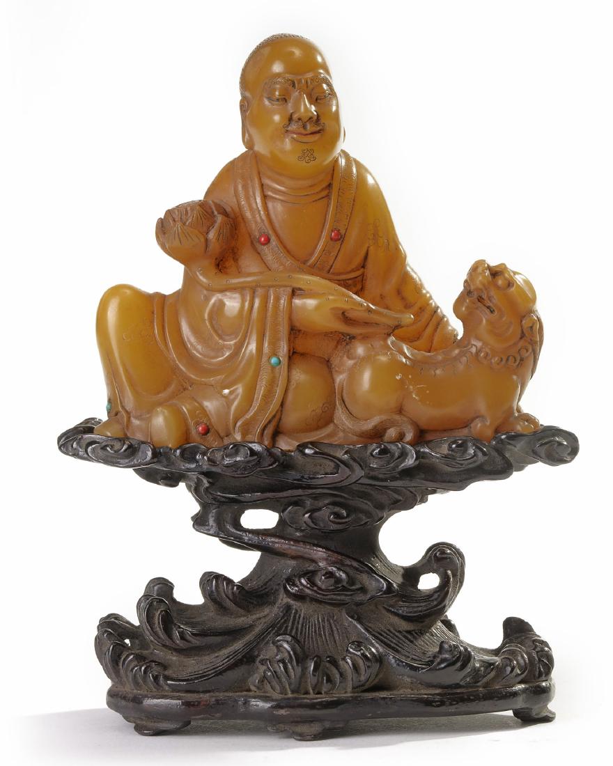 A Chinese soapstone carving of a Luohan: The Luohan is carved reclining holding a lotus stem in the crook of his right arm, accompanied by a recumbent Buddhist lion. The robe are decorated with incised flowers and various hardstone inlays. W