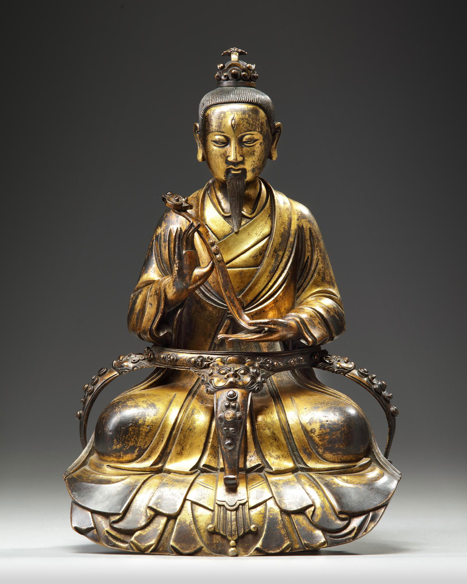 A CHINESE GILT BRONZE FIGURE OF A SCHOLAR: The figure is cast with a serene expression on his face, he wears scholar's cap and robes and holds a ruyi-sceptre in his hands. He is seated behind a platform raised on legs emanating from beast head
