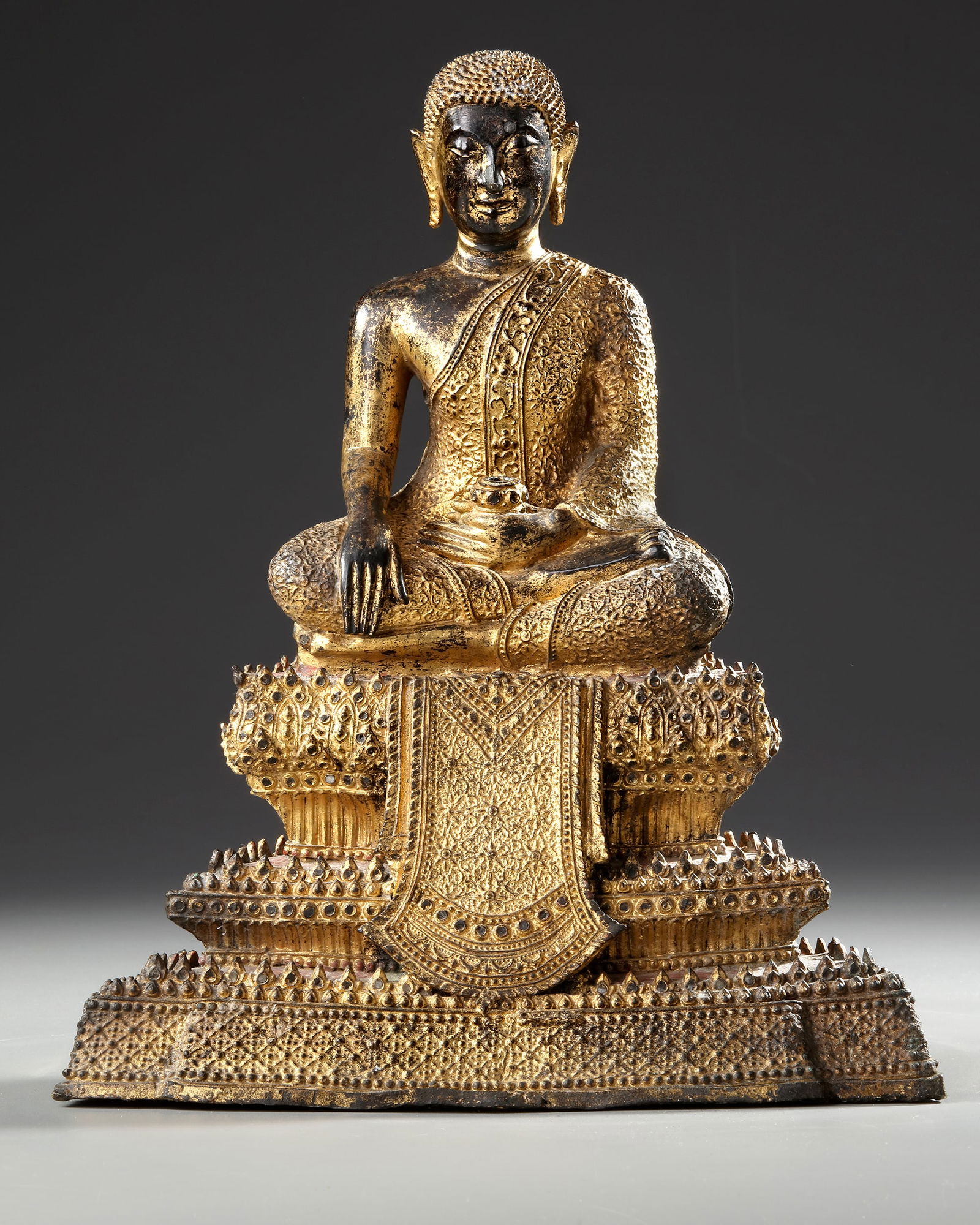 A GILT BRONZE SEATED BUDDHA FIGURE, THAILAND 19TH CENTURY: Cast seated in sattvasana on a shaped plinth, with his right hand in bhumisparsha mudra, his face in a serene expression of meditation, with arched brows and pendulous earlobes, his hair curled, with