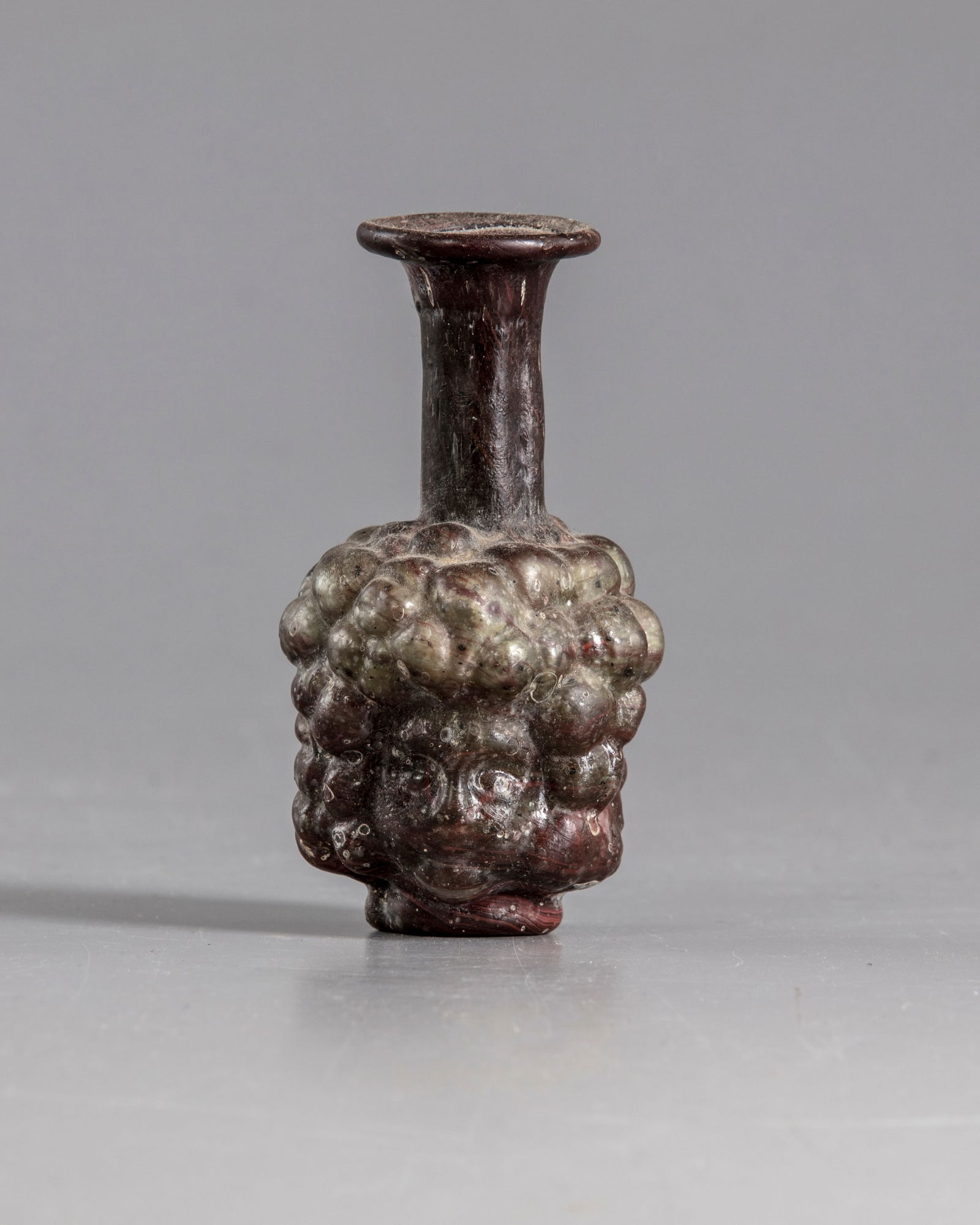 A ROMAN PURPLE GLASS HEAD FLASK, CIRCA 3RD CENTURY AD: A Roman purple glass head flask, mold-blown in two parts of heads. Flask with body in form of two human heads back to back, with short tapering cylindrical neck and everted rim, their curly hair repre