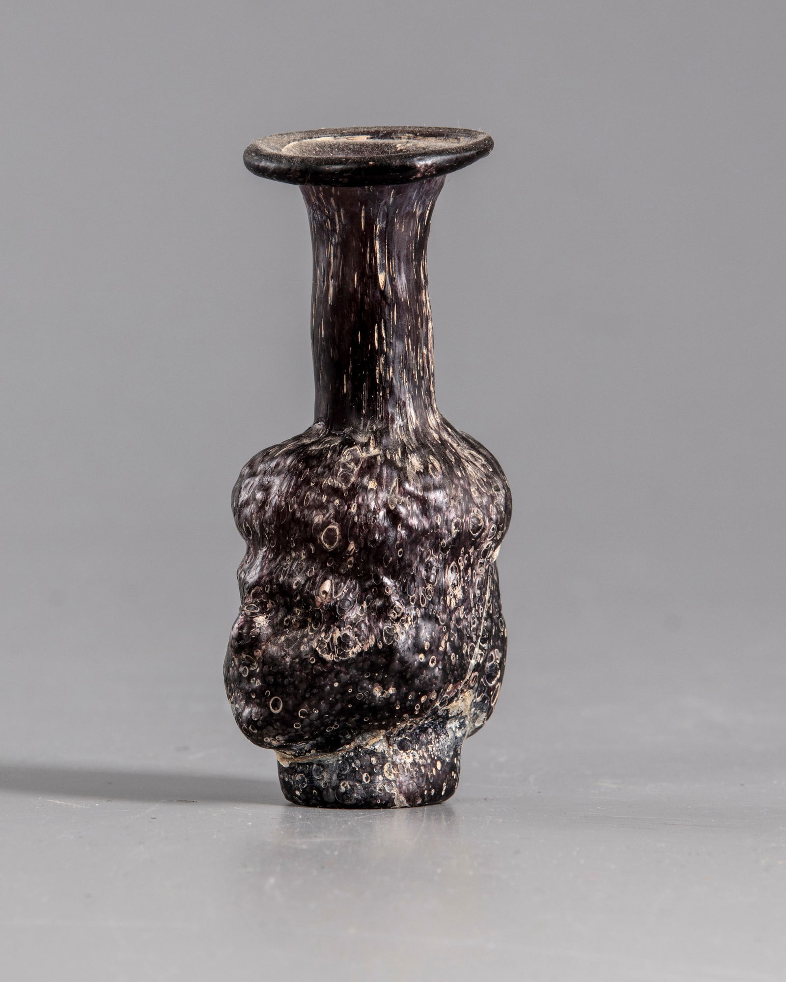 A ROMAN MOULDED PURPLE GLASS HEAD FLASK, CIRCA 3RD CENTURY AD: A Roman purple glass head flask, mold-blown in two parts of heads. Flask with body in form of two human heads back to back, with short tapering cylindrical neck and everted rim, with long hair indicat