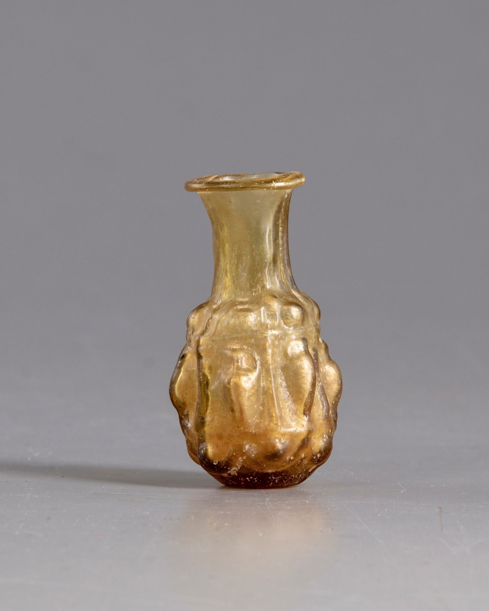 A ROMAN GLASS FLASK, CIRCA 3RD CENTURY AD: A Roman glass flask, the neck with flaring mouth and rounded inward-folder rim. the body decorated in relief with six vessels each set within a niche supported by columns, with a slightly flat base. H