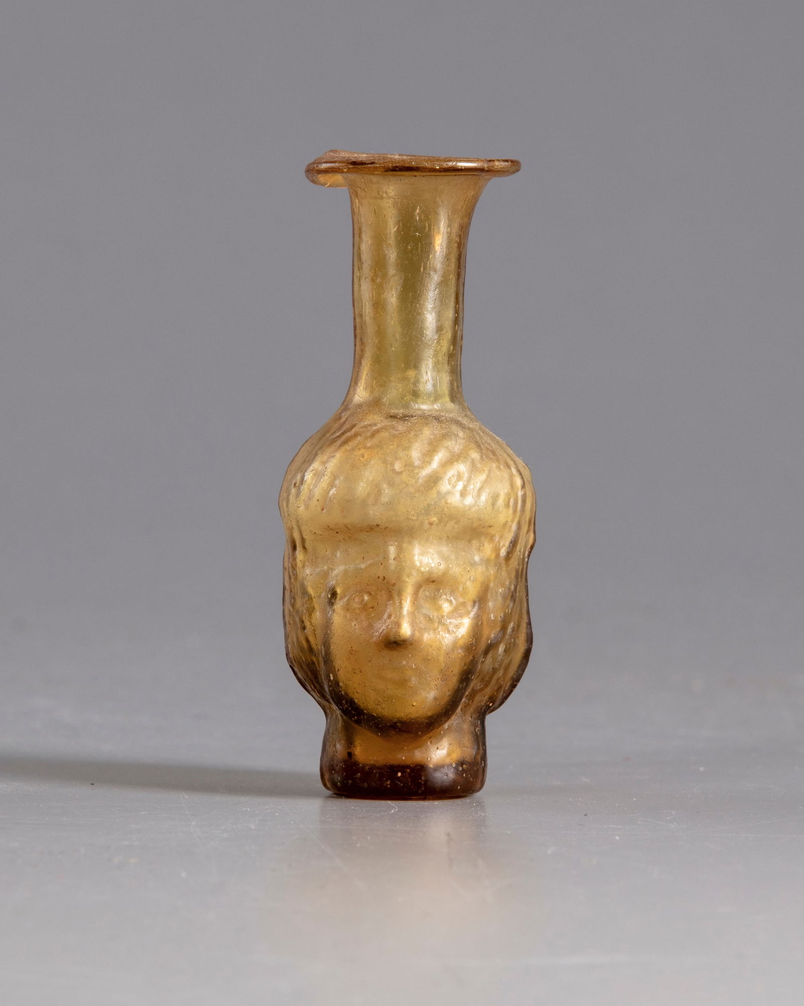A ROMAN HEAD GLASS FLASK , CIRCA 3RD CENTURY AD: A Roman glass flask , made of metallic yellow mould-blown glass. The vessel features a long tapering, cylindrical neck, a flaring rounded lip, and a rounded body resembling two identical faces of a Wo