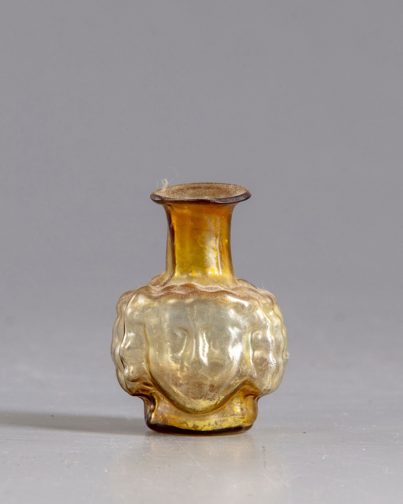 A ROMAN HEAD GLASS FLASK, CIRCA 3RD CENTURY AD: A Roman glass flask , made of metallic yellow mould-blown glass. The vessel features a short, cylindrical neck; a flaring rounded lip; and a rounded body resembling two identical faces of youthful che