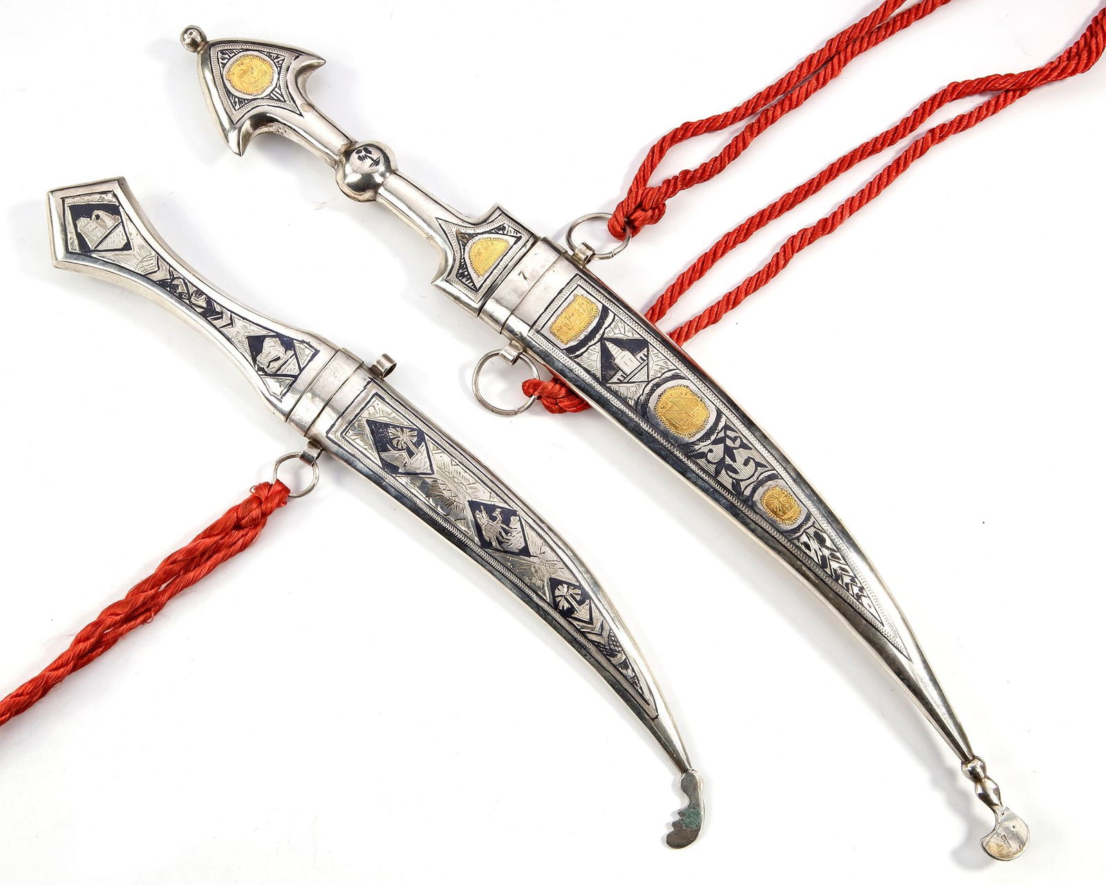 TWO SILVER DAGGERS – IRAQ, 20TH CENTURY: A matched pair of Iraqi daggers, each with finely crafted silver sheaths decorated with black and gold inlays of geometric and symbolic motifs. The blades are slightly curved, housed in polished silve