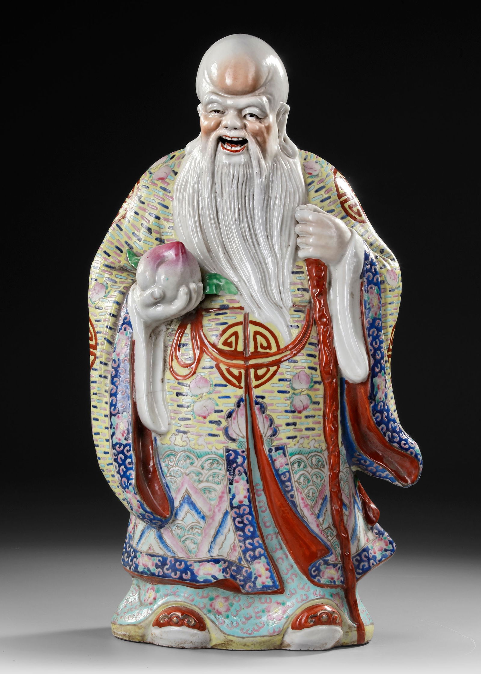 A LARGE CHINESE FAMILLE ROSE FIGURE, 19TH-20TH CENTURY: A finely modeled and brightly enameled porcelain figure of Shoulao, the Chinese god of longevity, depicted holding a peach of immortality and a staff. His flowing robes are richly decorated with auspi