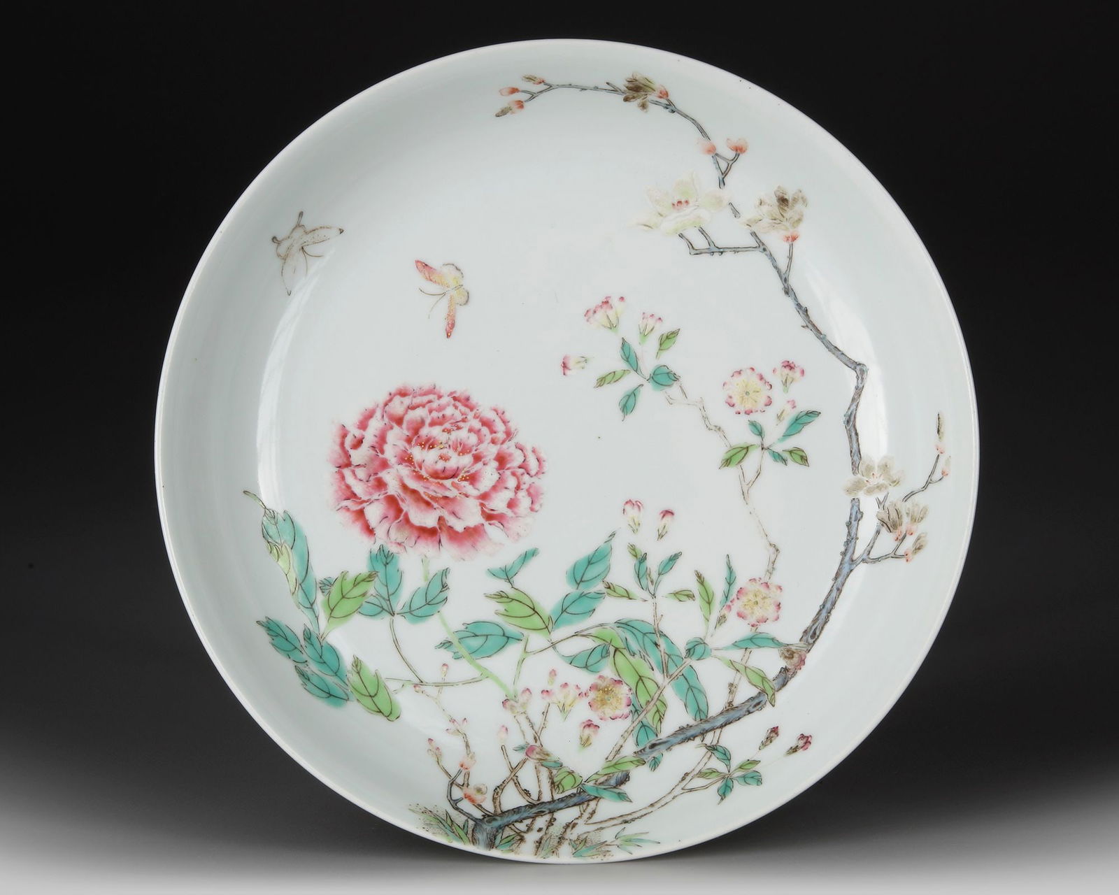 A CHINESE FAMILLE ROSE 'FLORAL DISH, 19TH CENTURY: A famille rose porcelain dish delicately painted with a vibrant peony bloom, blossoming branches, and fluttering insects. The reverse bears a six-character Qianlong mark. Diameter 20 cm. 
