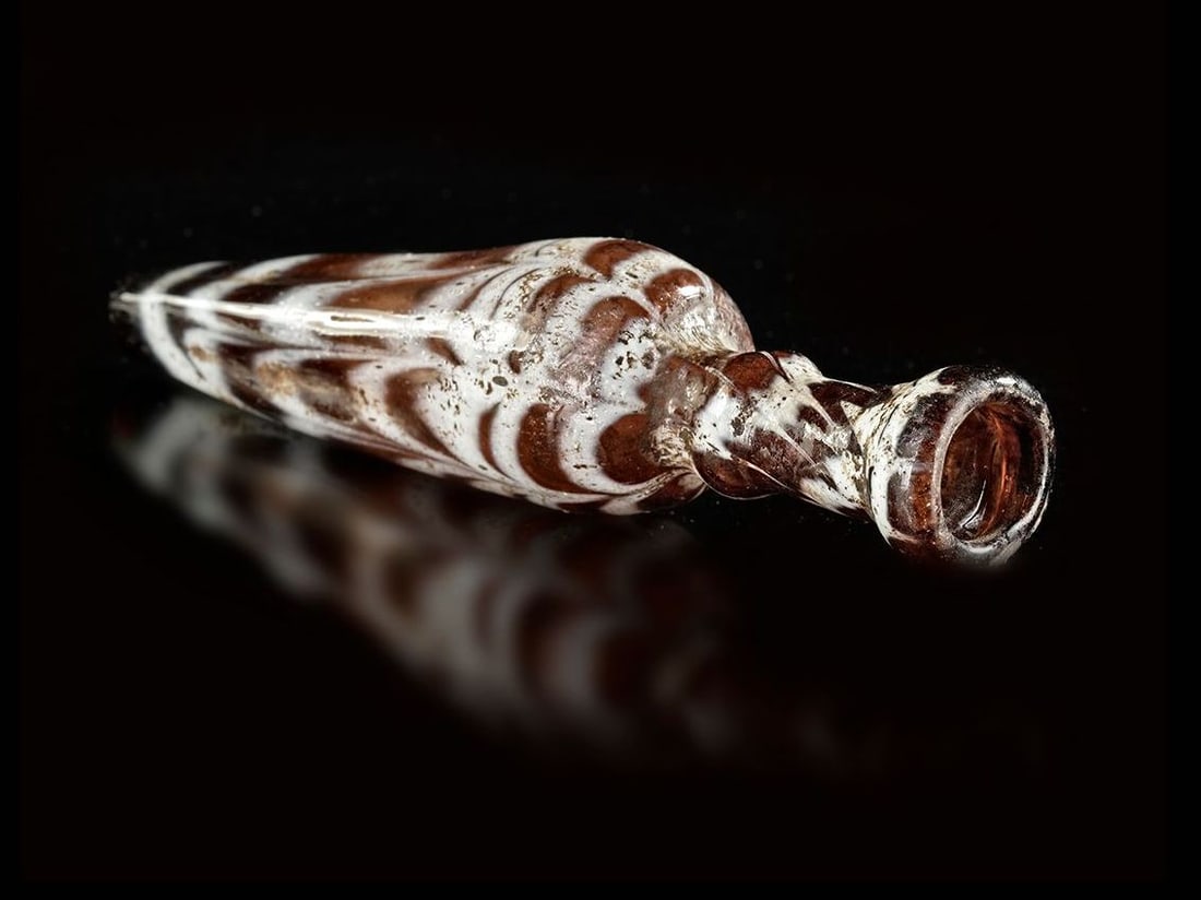A SMALL KOHL FLASK, SYRIA 12TH-13TH CENTURY: Spindle-shaped flask in translucent purple-brown glass marbled with white threads, its cylindrical neck with swollen base and slightly hemmed lip. Little missing at the base. Similar flasks can be