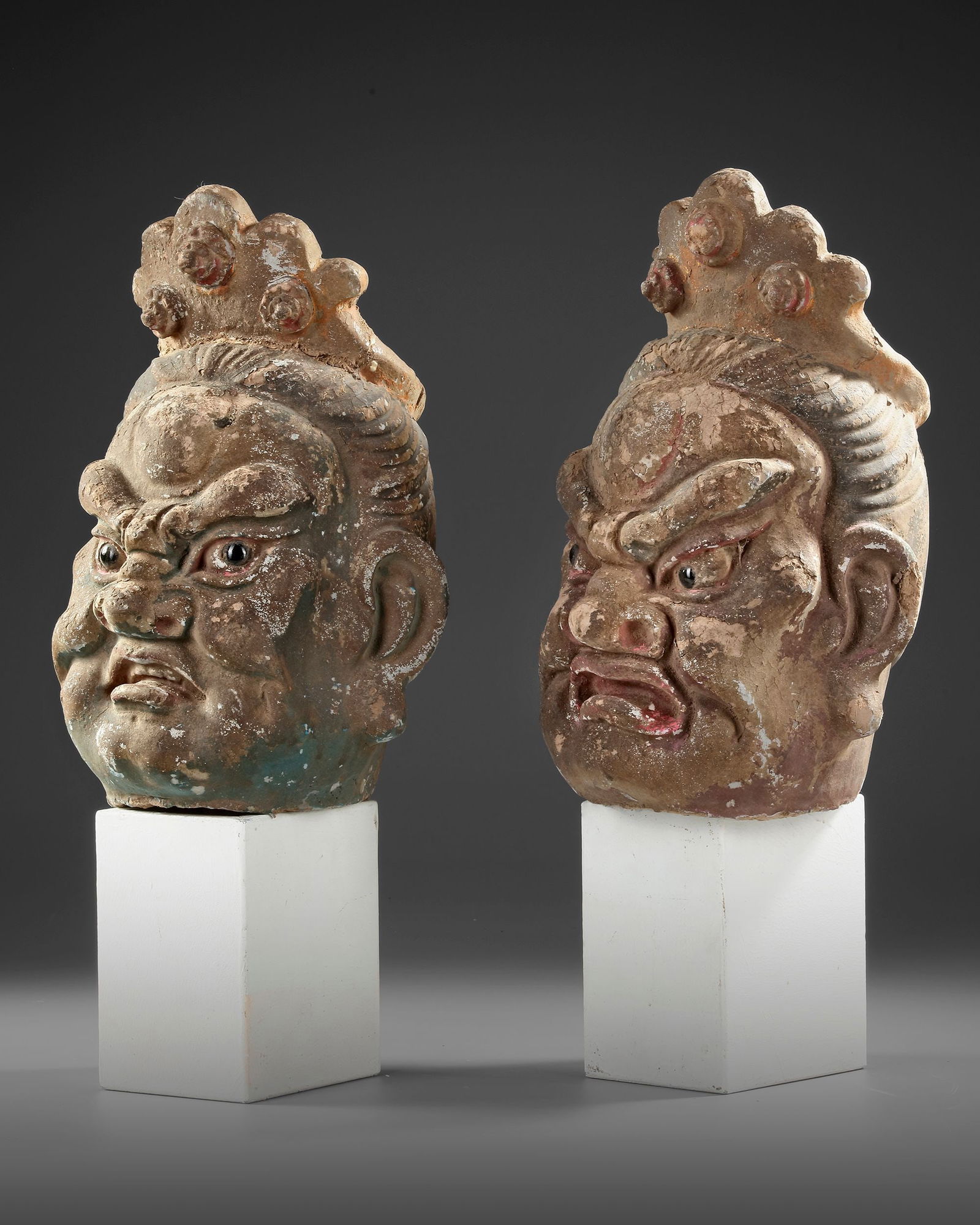 TWO CHINESE POLYCHROME WOOD HEADS, MING DYNASTY OR LATER: A pair of Chinese carved and polychrome-painted wooden heads, each depicting a fierce guardian figure with expressive features and ornate crowns. The remnants of original pigments highlight the detail