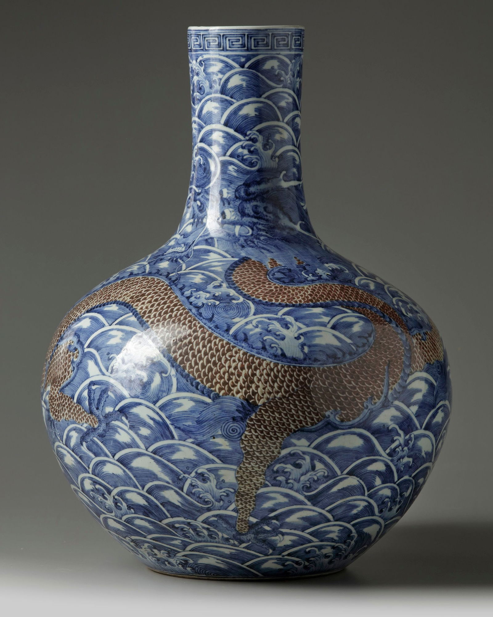 A LARGE CHINESE UNDERGLAZE COPPER RED AND BLUE AND WHITE 'DRAGON' VASE, QING DYNASTY (1644-1911): The vase is decorated with a bold writhing dragon with scaled in underglaze copper red which winds itself around the body and up the neck, all on a ground of densely foaming waves. The rim is decorate