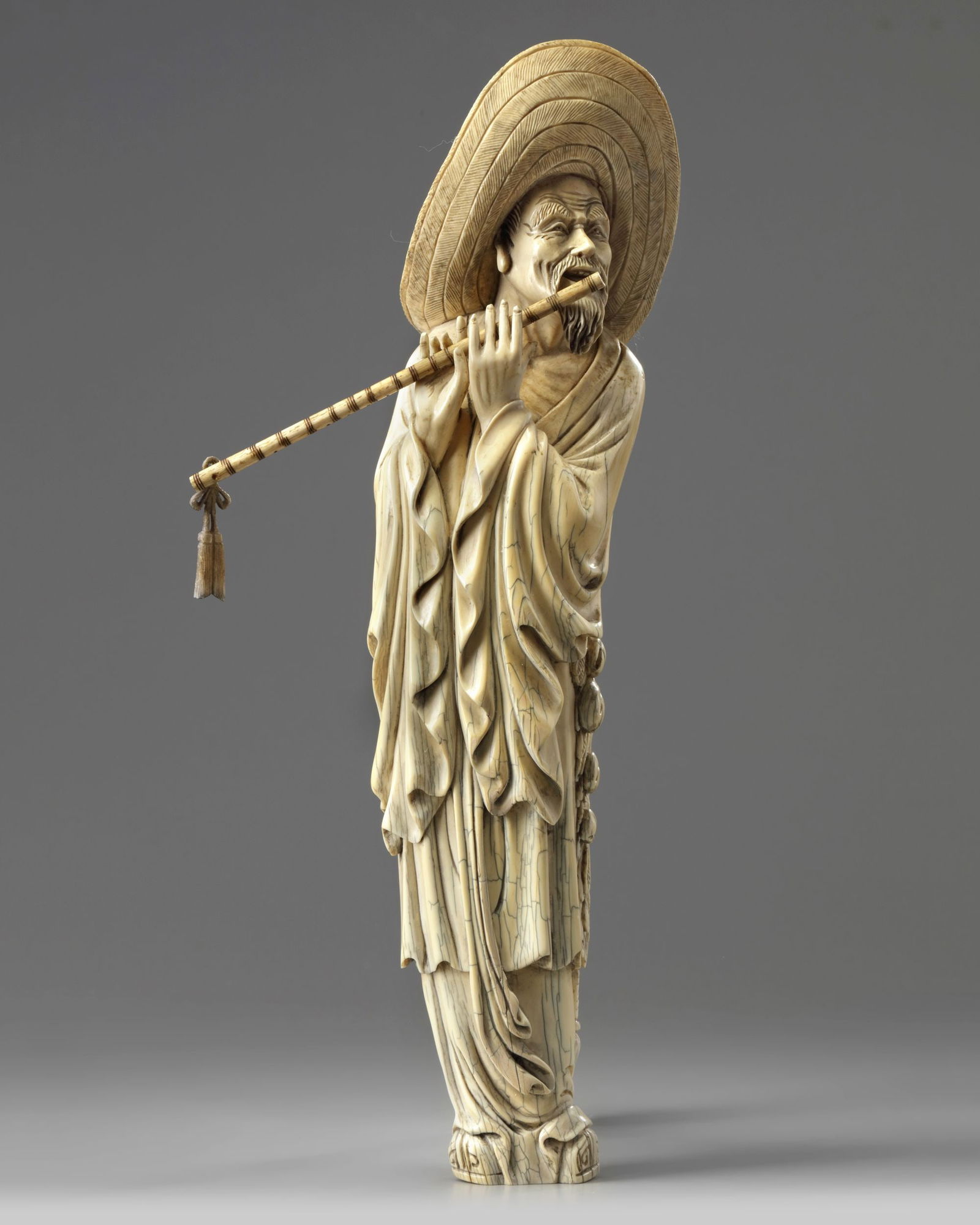 A CHINESE CARVED BONE MUSICIAN, 19TH-20TH CENTURY: The figure is carved wearing a large wide-brimmed hat and long robes. He is carved smiling with two hands playing a detachable flute. Height 35 cm.
