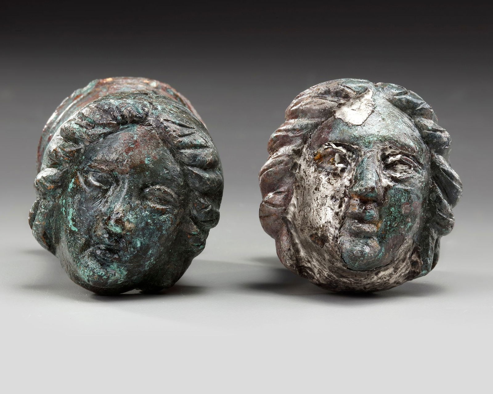 A PAIR OF ROMAN BRONZE AND SILVERED 'FACE OF FEMALE' TERMINALS, CIRCA 2ND-3RD CENTURY AD: A cast bronze of two female head terminals probably for a chair or furniture. Left to right: 6.5 cm., 5.5 cm. length.PROVENANCE Private collection, Germany