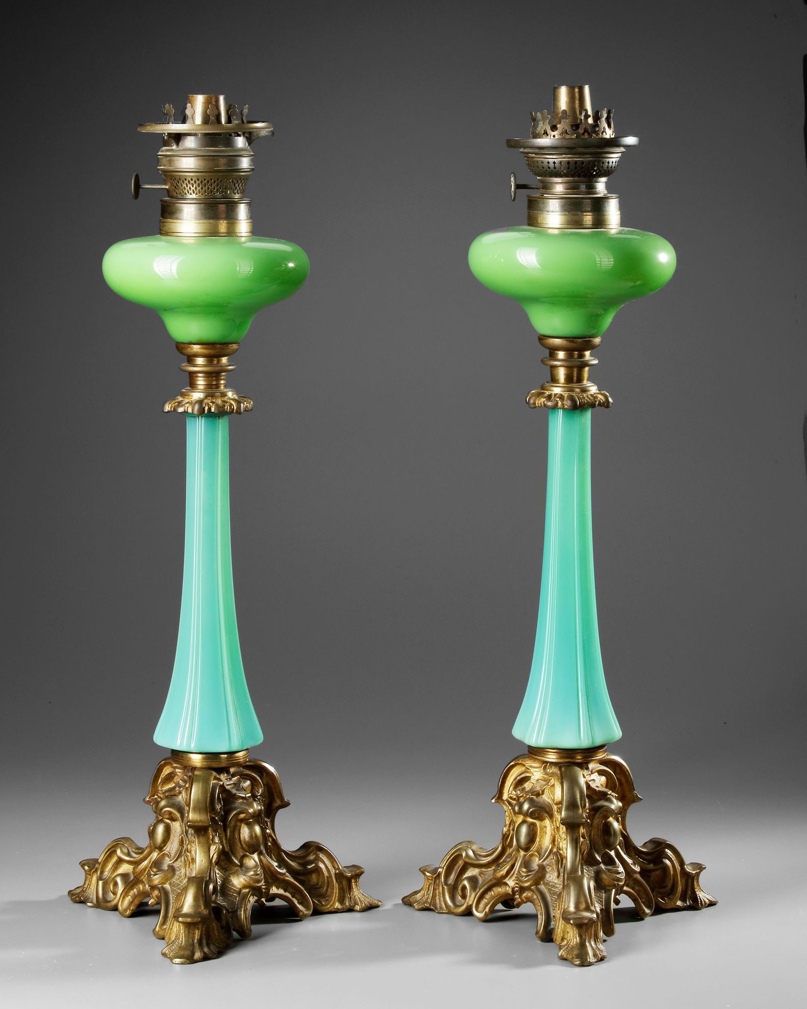 A PAIR OF OILLAMPS, 19TH CENTURY: A pair of 19th-century oil lamps featuring ornate gilt bronze rococo-style bases and slender turquoise glass stems. Topped with green glass reservoirs, these lamps combine elegance and craftsmanship,