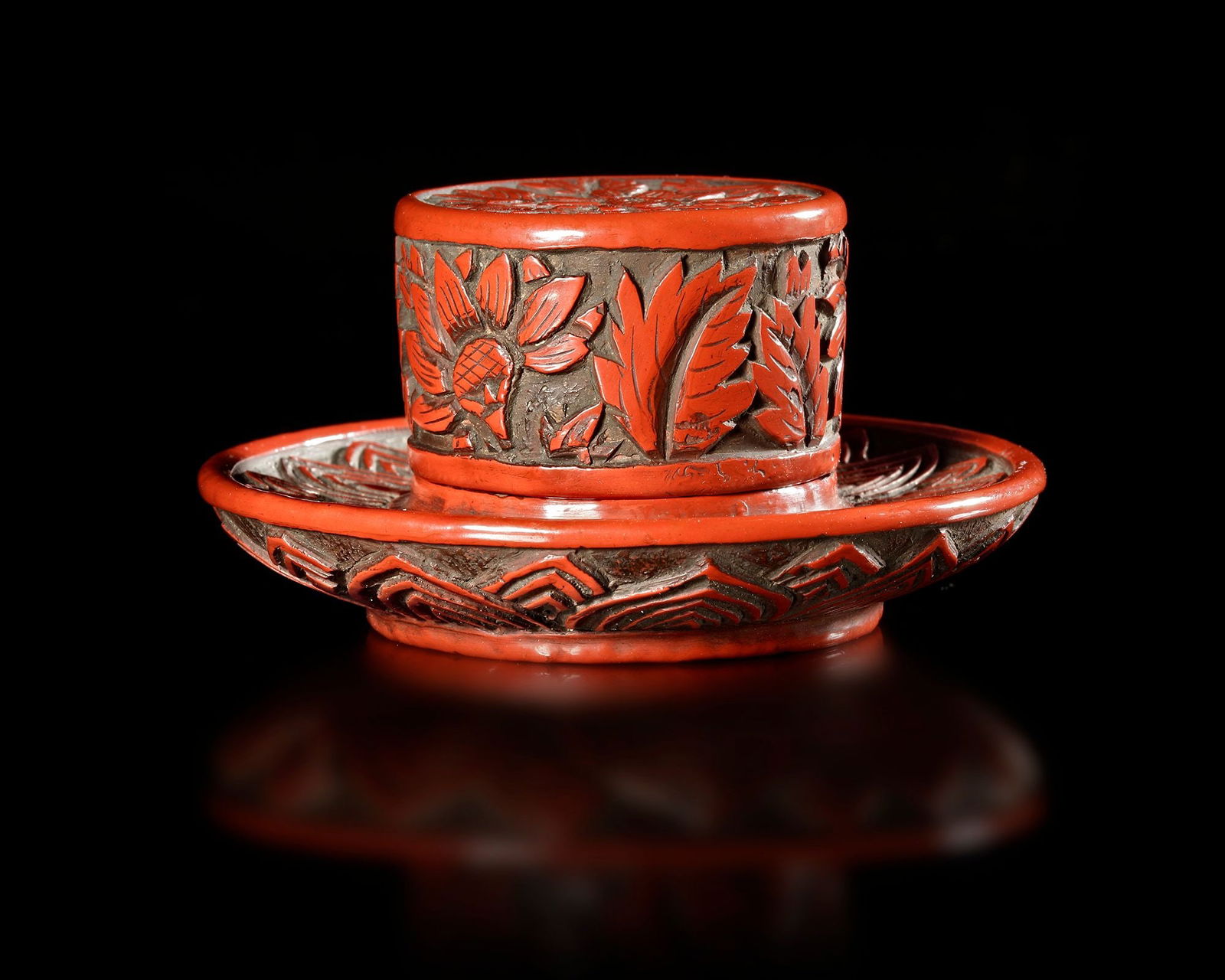 A RED LACQUER INK CONTAINER, CHINA, MING DYNASTY (1368-1644): A finely carved red lacquer ink container.Decorated with floral motifs in relief. The cylindrical container sits on a matching lacquer stand, both richly ornamented with traditional Chinese patterns.