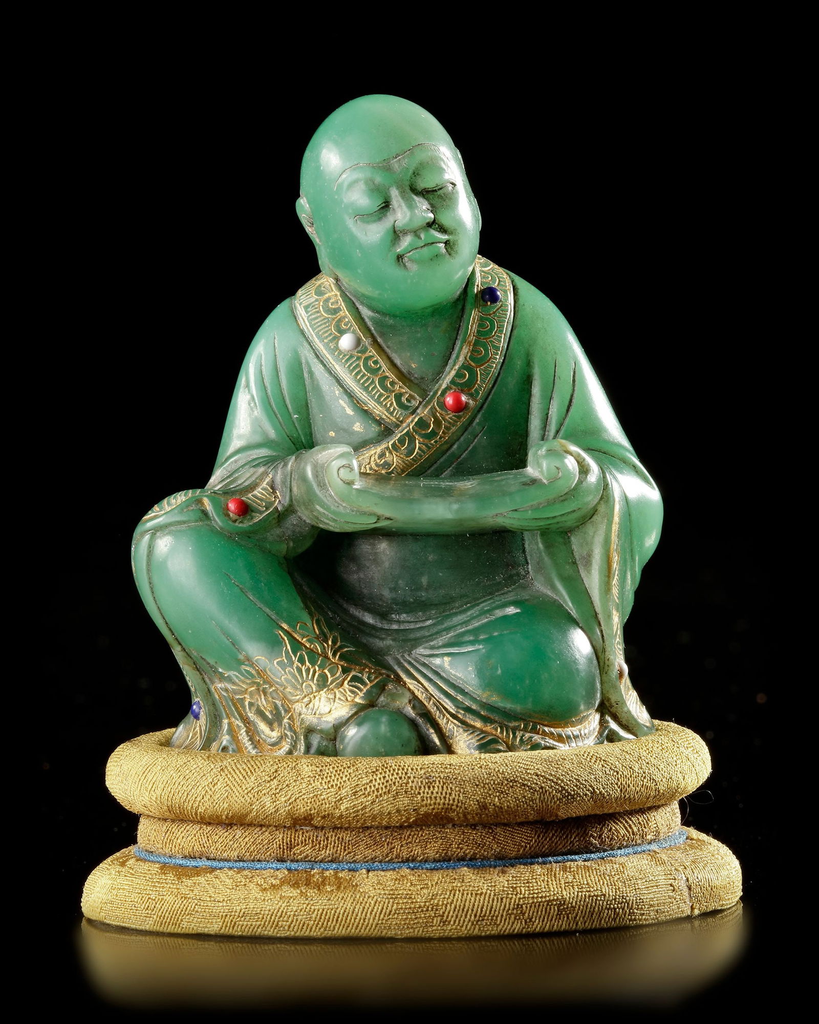 A LUOHAN FIGURE, CHINA, QING DYNASTY (1644â€“1912): Green Steatite Finely carved figure represents a Luohan (Arhat), a revered Buddhist disciple who has attained enlightenment. The figure is shown seated in a contemplative posture, holding a scroll whi