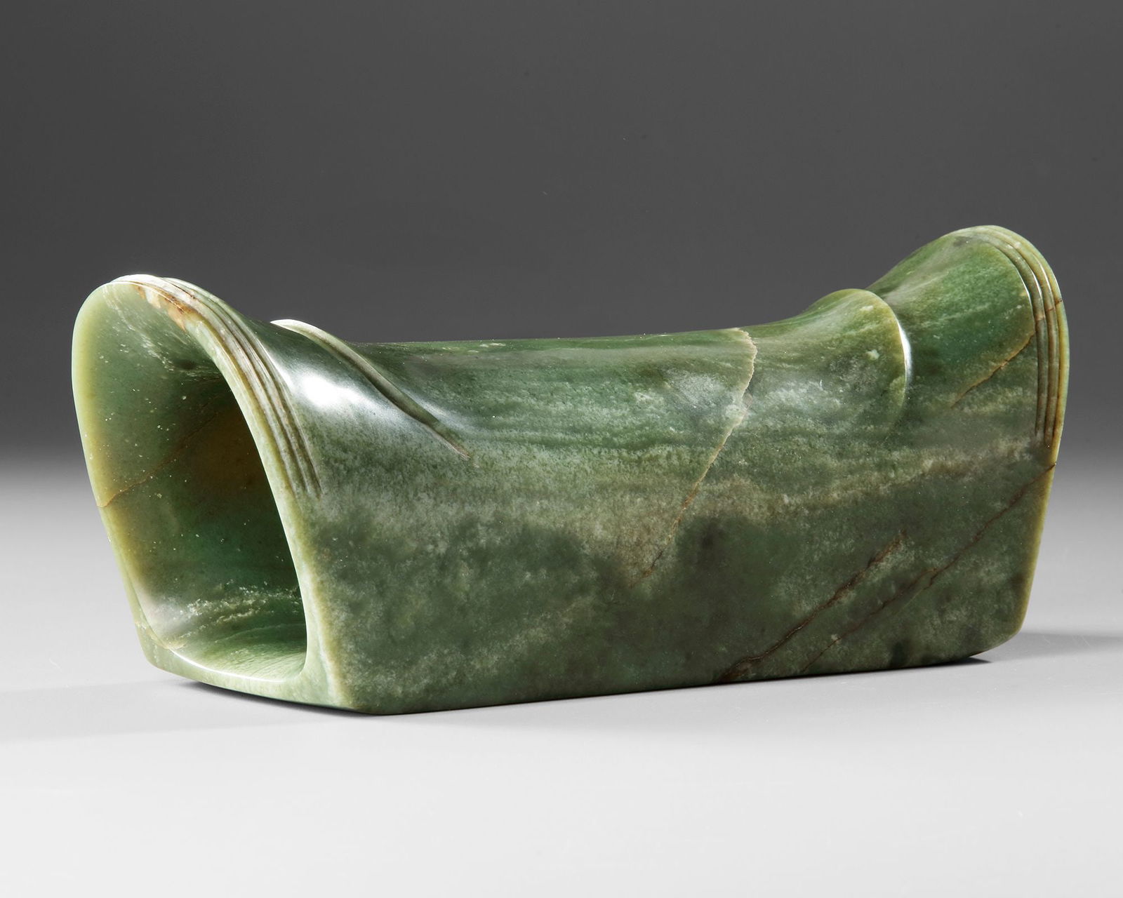 A JADE HEADREST, CHINA, SONG DYNASTY: A finely carved headrest made from mottled green jade, dating to the Song Dynasty. The piece displays a smooth, gently curved form designed for comfort and support, with natural marbling and tonal var