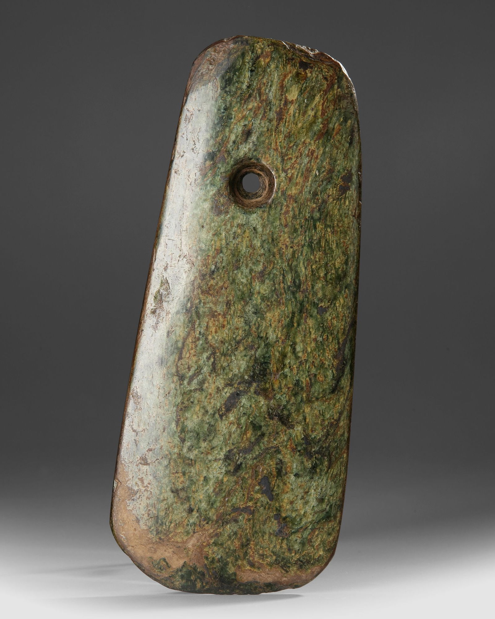 A NEOLITHIC GREEN JADE AXE, CHINA: Large ceremonial axe carved from mottled green jade, dating to the Neolithic period. The elongated rectangular form features smoothly polished surfaces and a drilled suspension hole, likely used for h