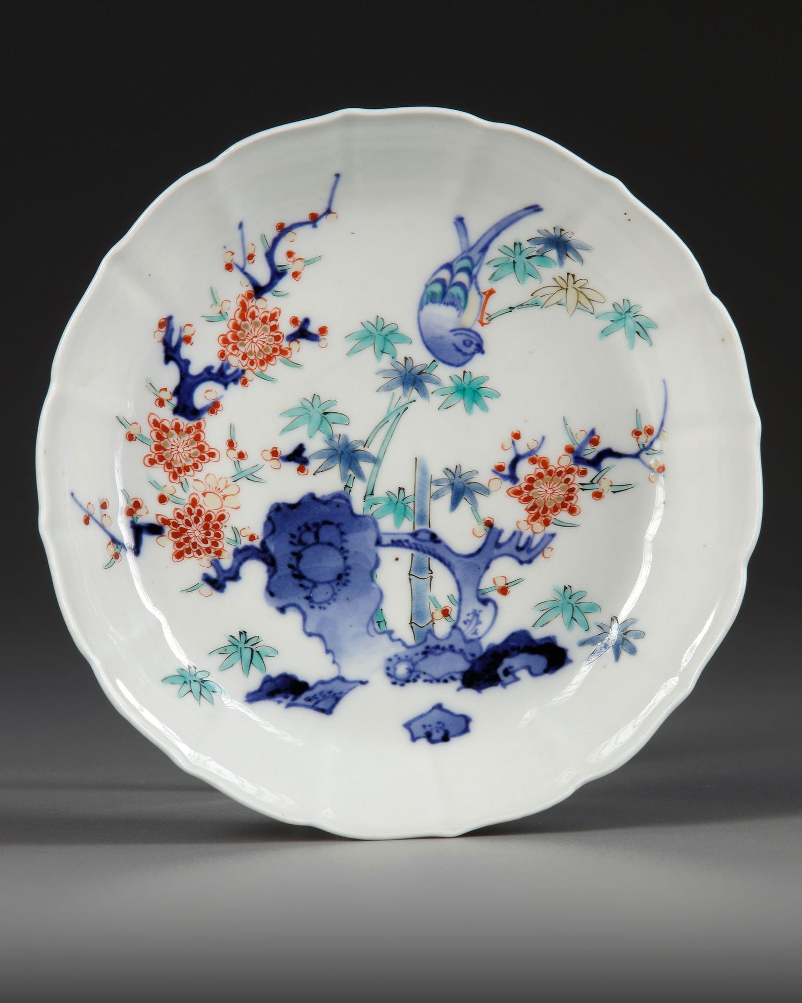 A JAPANESE PORCELAIN KAKIEMON DISH, EDO PERIOD, 18TH CENTURY: A Japanese porcelain dish with flutted sides and barbed rim, brightly enameled in blue, turquoise and red enamels with the Kakiemon pattern of a bird perched on a tree, surrounded by pine, prunus and