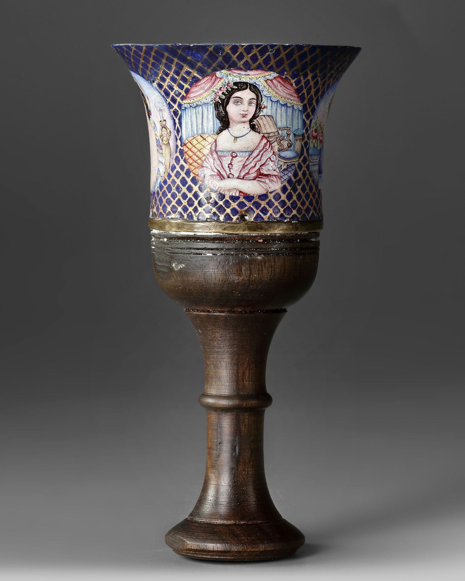 AN OTTMAN POLYCHROME ENAMELLED GHALIAN CUP,19TH CENTURY: Ottoman polychrome enamelled ghalian cup with painted figural scenes set against a deep blue ground with gilt lattice decoration, mounted on a turned wooden stem. H. 17 cm.