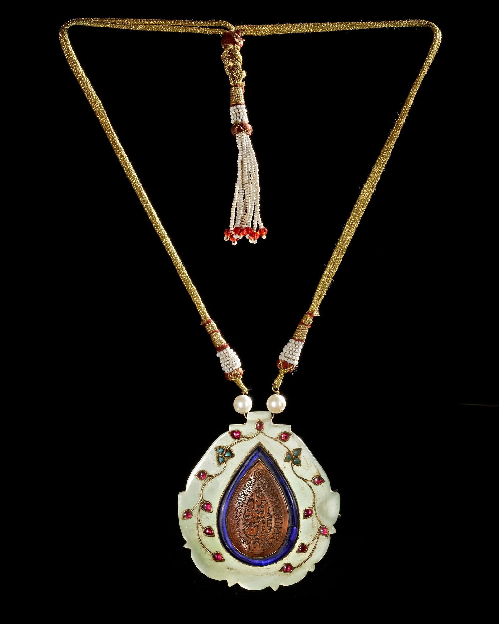 NEPHRITE JADE AMULET PENDANT, 20TH CENTURY: An Indo-Persian amulet pendant carved from celadon nephrite jade, set with a carnelian medallion engraved with a Qurâ€™anic verse. The medallion is framed by a dark blue glass border with flor